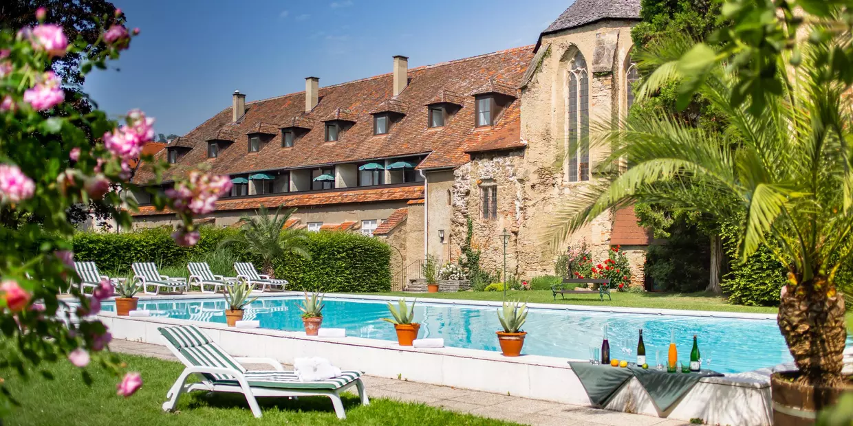 Sunbathing lawns with sunbeds, outdoor pool and, in the background, the old convent walls of today’s Schlosshotel Richard Löwenherz beneath a radiantly blue sky