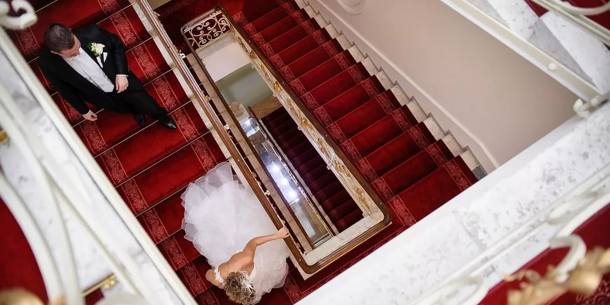 Elegantes, mit rotem Teppich ausgelegtes Treppenhaus im Hotel Elizabeth in Trencin, auf welcher ein Brautpaar hinabschreitet (c) Foto Hotel Elizabeth