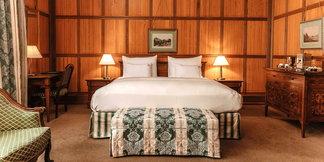 Double room (c) Hotel König von Ungarn In front of a wood-paneled wall stands a white-upholstered double bed with an elegant upholstered bench at the foot of the bed; to the left of the bed is an antique desk, and to the right is a dresser with a coffee maker (c) Photo: Hotel König von Ungarn