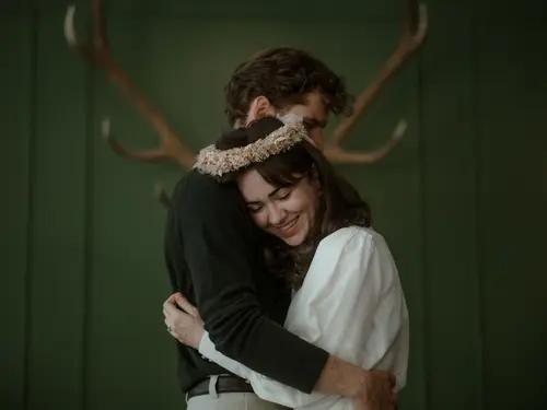 Wedding at the Schlosswirt zu Anif (c) Selina Schöberl A wedding couple embracing closely at the Schlosswirt in Anif in front of a dark green wall featuring a deer trophy; the bride is dressed in white with a white floral wreath in her hair (c) Photo by Selina Schöberl