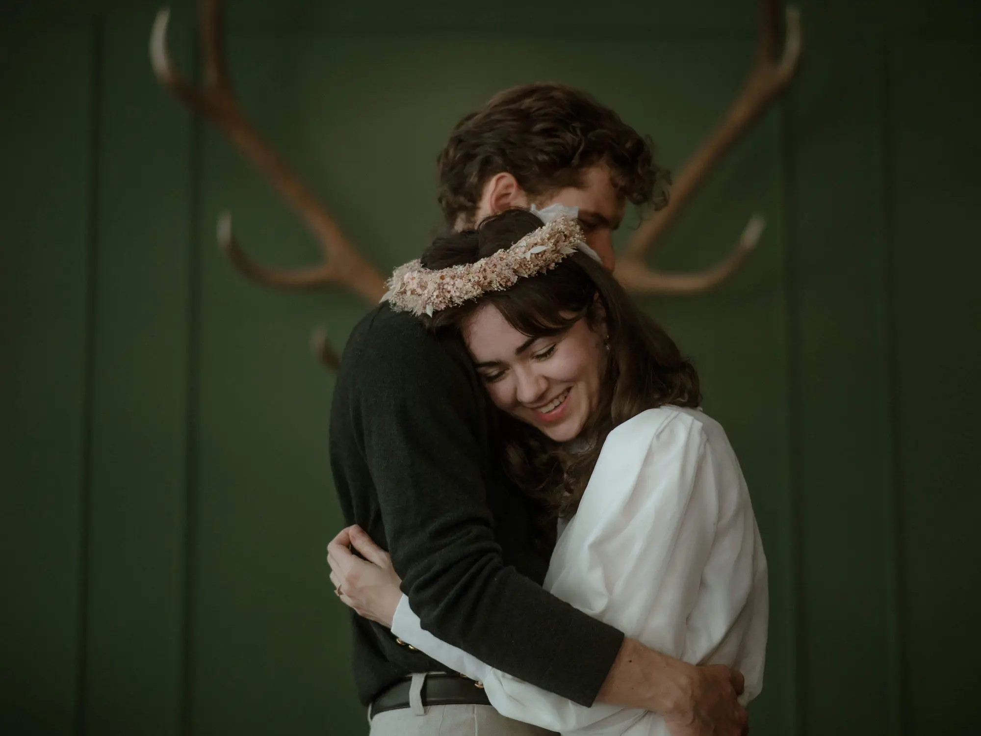 Eng umschlungenes Hochzeitspaar im Schlosswirt zu Anif vor einer dunkelgrünen Wand mit einer Hirschtrophäe, die Braut in Weiss mit einem weissen Blumenkranz im Haar (c) Foto Selina Schöberl