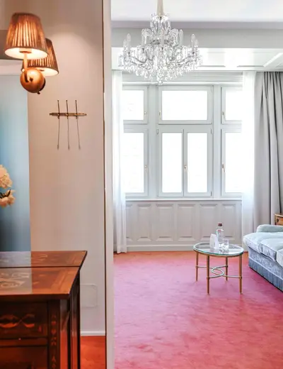 Junior suite (c) Hotel König von Ungarn View into a junior suite with an antique side table in the entryway and a view toward the window, featuring a couch on the right side of the image and a crystal chandelier on the ceiling (c) Photo: Hotel König von Ungarn