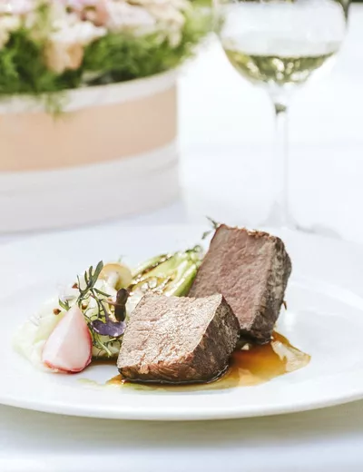Meat dish on a white plate with floral decoration and a glass of wine at Restaurant Schlosshotel Szidonia in Röjtökmuzsaj (c) photo Fenyertek Studio Kft.