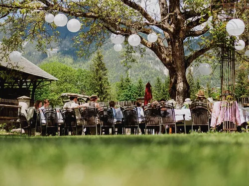 Langer Tisch unter einem alten Baum mit Gästen im Garten vom Hotel Schloss Lerchenhof in Hermagor, Kärnten